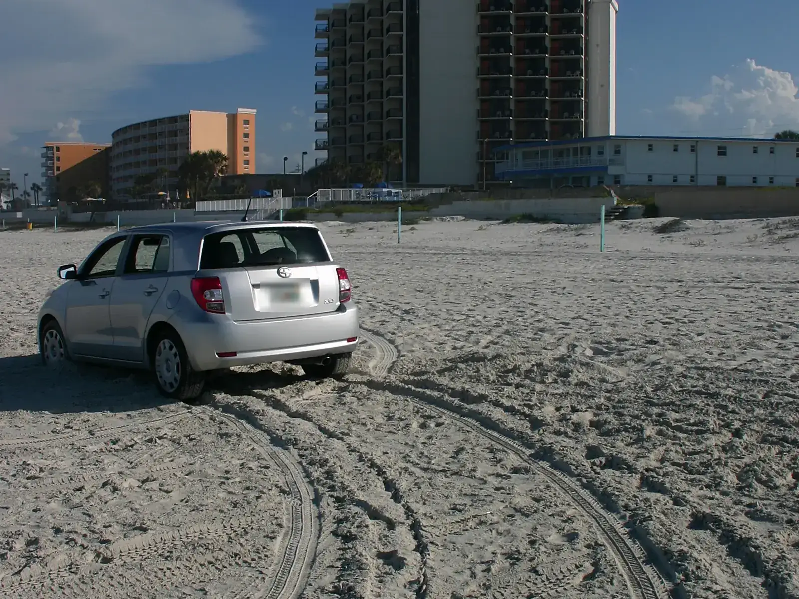 Silver hatchback on packed beach sand with tire tracks and beachfront buildings behind, on the sand at Daytona Beach
